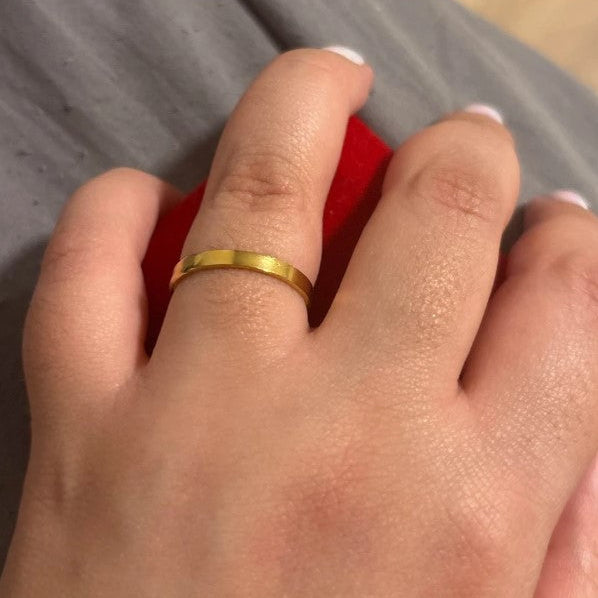 A close-up of a person's hand wearing a simple, polished yellow gold band ring on the ring finger, resting on a red surface with gray fabric in the background.
