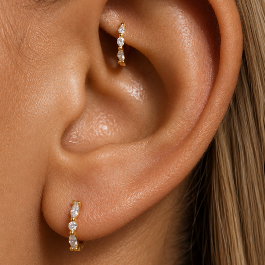 Close-up of a person's ear wearing two small gold ear cuffs with clear stones, shown on skin with blonde hair visible.