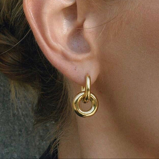 A close-up of a person's ear wearing a small gold hoop earring with a larger circular drop hoop attached below.