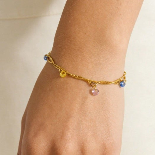 A delicate gold vine bracelet with small dangling charms, including blue and yellow beads and a pink teardrop charm, shown worn on a person's wrist against a soft cream background.