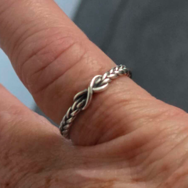 A close-up of a braided silver ring with an infinity symbol design worn on a finger.