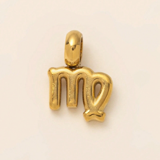 A polished gold pendant shaped like the astrological symbol for Virgo, displayed on a clean, light beige background.