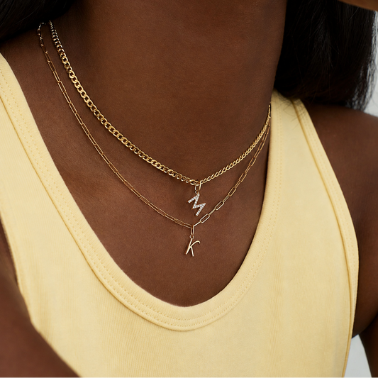 A close-up of a person wearing layered necklaces over a pale yellow ribbed tank top. The necklaces include a gold chain, a fine gold chain, and a silver chain with a dangling initial pendant.