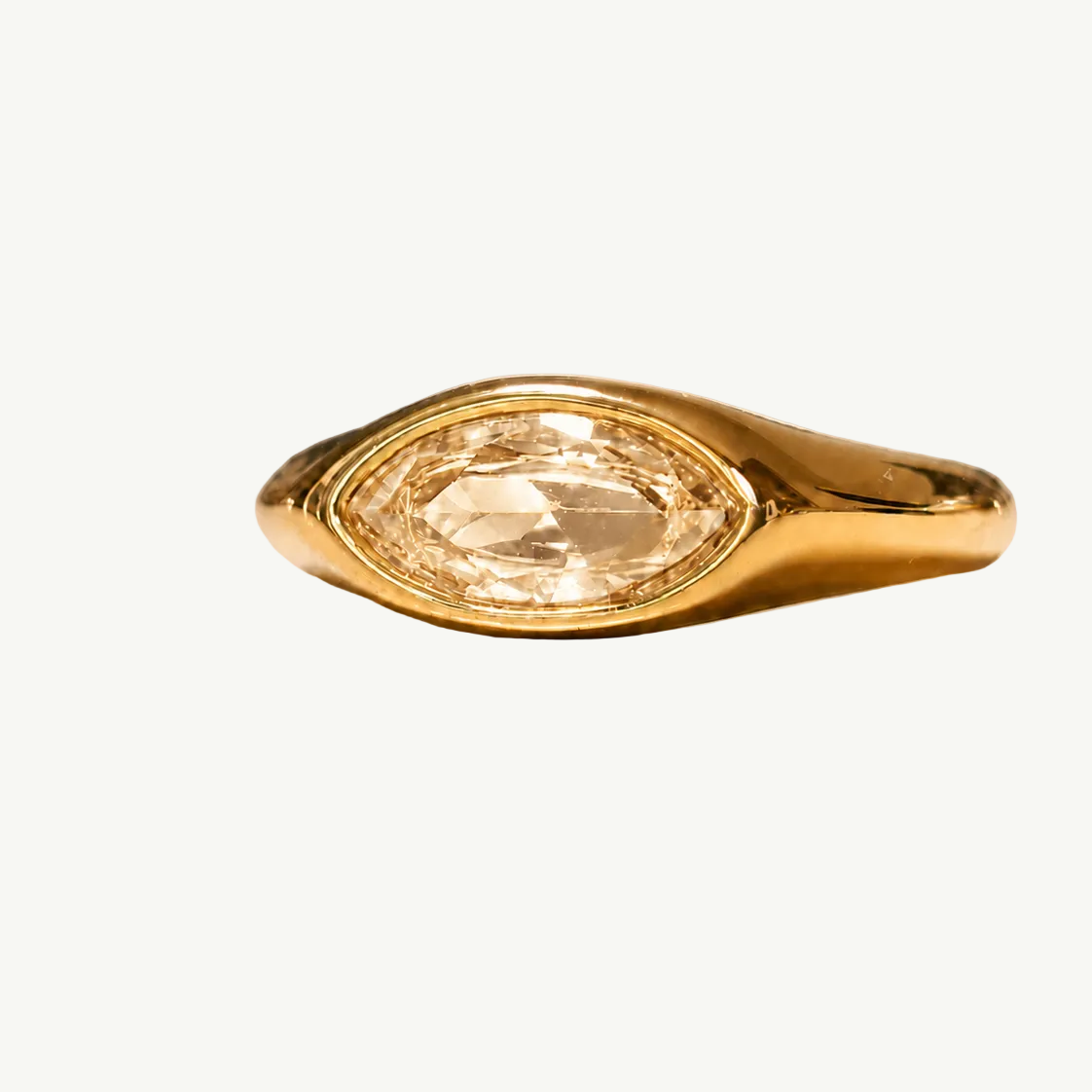 A gold ring with a marquise-cut clear gemstone, displayed on a warm beige reflective surface.