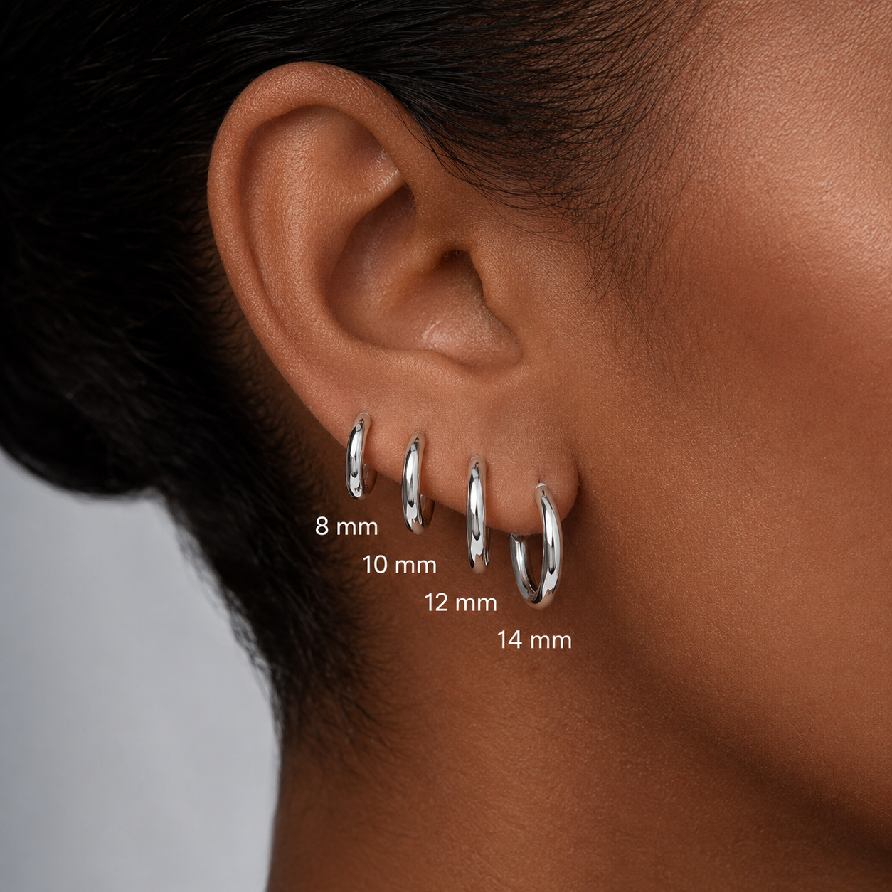 Close-up of a person's ear wearing four silver hoop earrings in different sizes labeled 8 mm, 10 mm, 12 mm, and 14 mm.