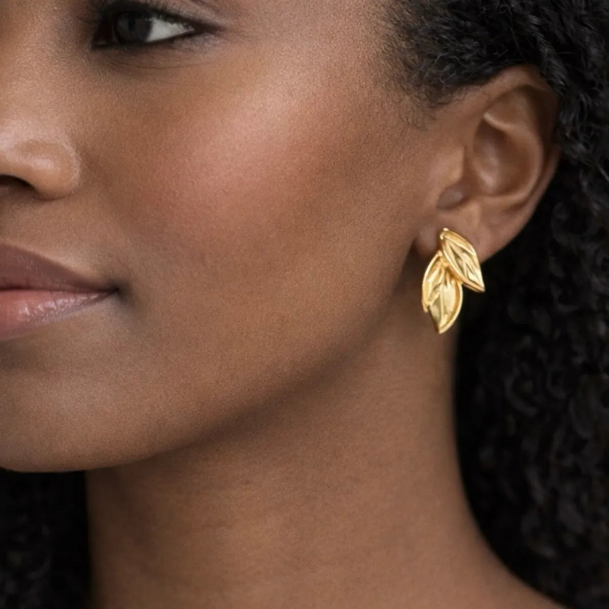A close-up of a woman wearing a gold leaf-shaped stud earring, showing the earring on her ear against her dark hair and skin.