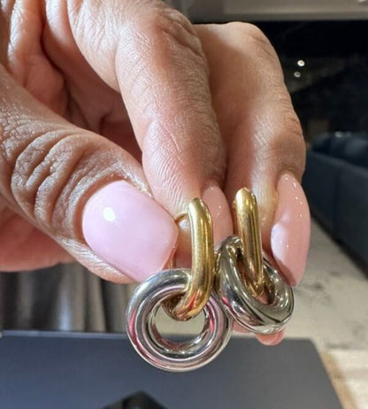 A close-up of a hand with light pink manicured nails holding a pair of gold and silver hoop earrings.