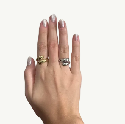 A close-up of a hand wearing two wrap-style rings, one in polished gold and one in polished silver, against a neutral indoor background.