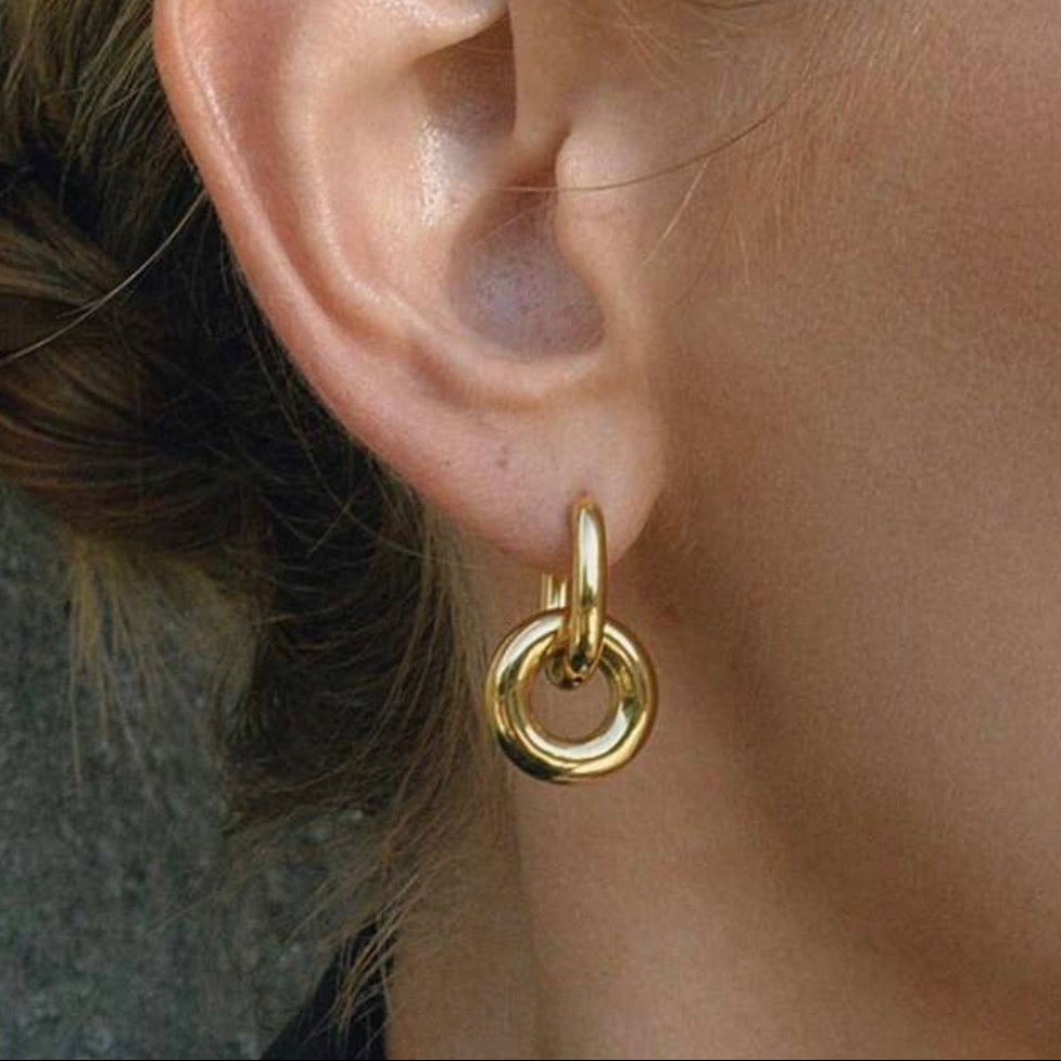 A close-up of a person's ear wearing a small gold hoop earring with a larger circular drop hoop attached below.