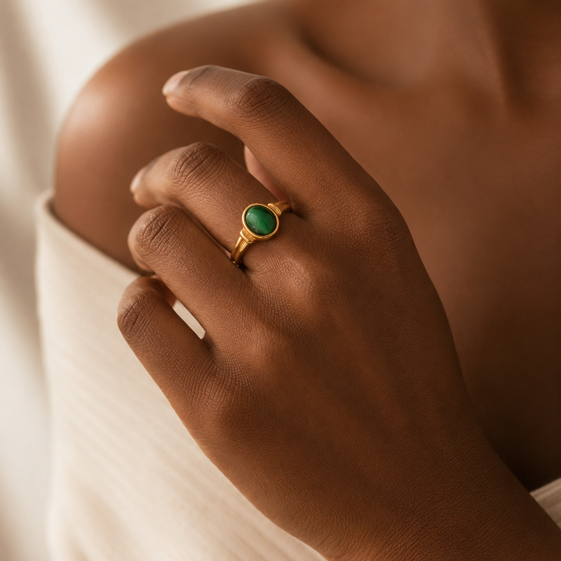 A close-up of a person's hand wearing a gold ring with a round green stone, set against a soft cream-colored ribbed top and a warm neutral background.