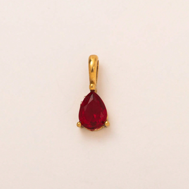 A gold pendant featuring a teardrop-shaped red gemstone in a prong setting, photographed against a plain off-white background.