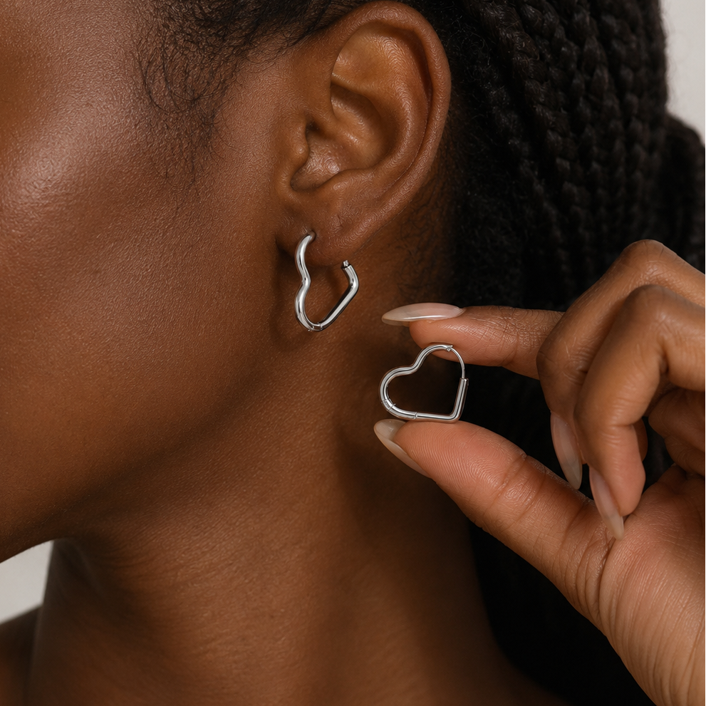 A close-up lifestyle shot of a woman wearing a silver wavy hoop earring, with her hand holding a matching silver heart-shaped hoop earring against a neutral light background.