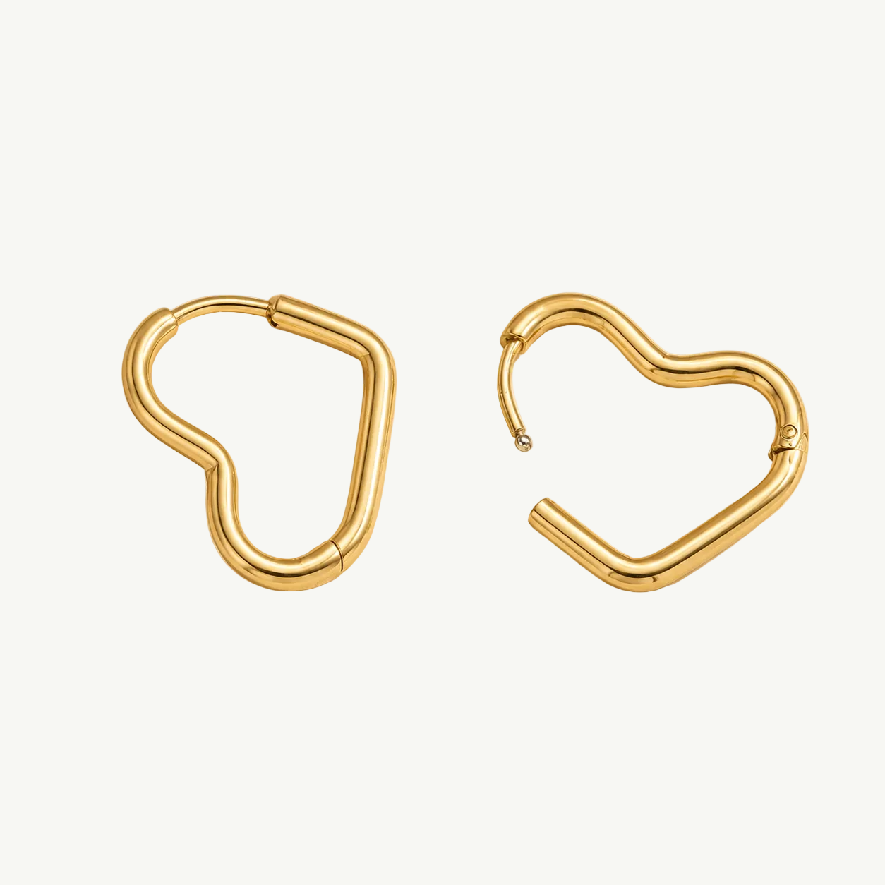 A pair of gold-plated heart-shaped hoop earrings shown on a warm beige background. One earring is closed, and the other is open, displaying its hinge mechanism.