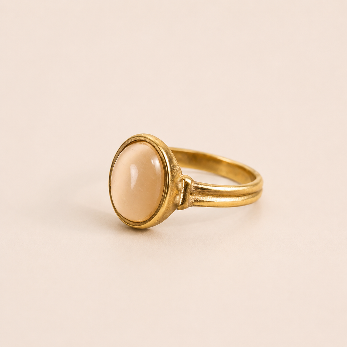 A gold ring with a smooth, oval-shaped pale peach cabochon stone, set against a solid light beige background.