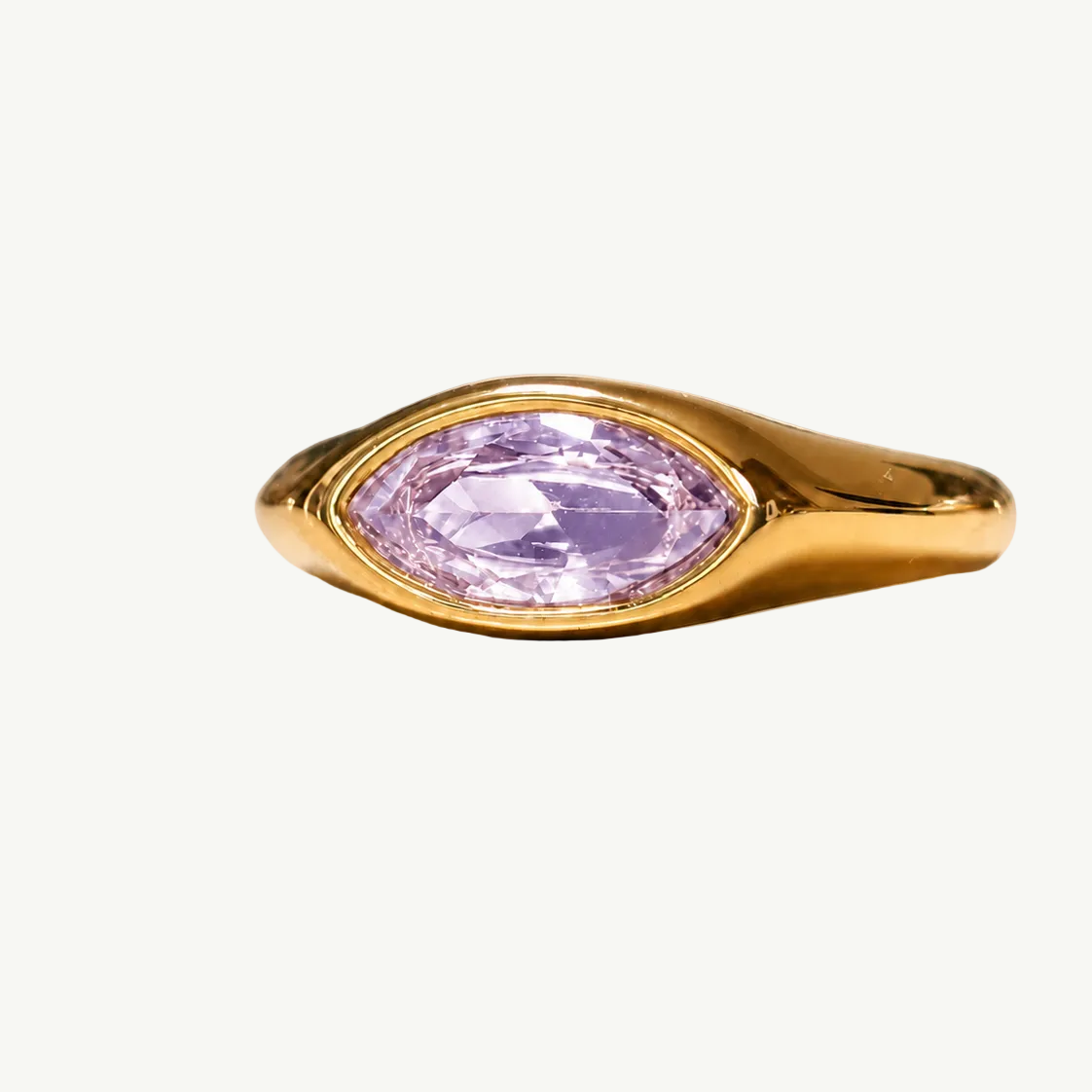 A gold ring featuring a marquise-cut light purple gemstone, displayed on a soft beige surface with a soft reflection.