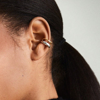 A close-up side profile of a person wearing a double stacked hoop earring in gold and silver over a black ear cuff, with dark hair tied back in a ponytail.