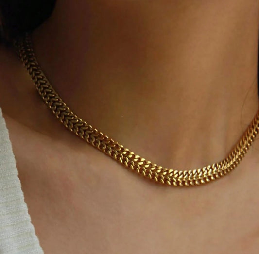 A close-up of a gold chain choker necklace worn on a person's neck, with a white ribbed top visible at the side.