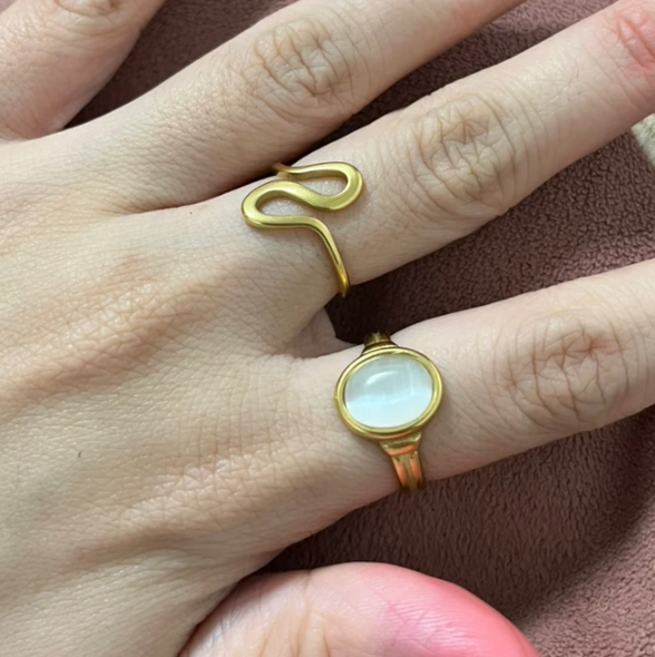A close-up of a hand wearing two gold rings: one is a snake-shaped ring on the middle finger, and the other is a gold ring with an oval milky white stone on the ring finger, set against a brown textured background.