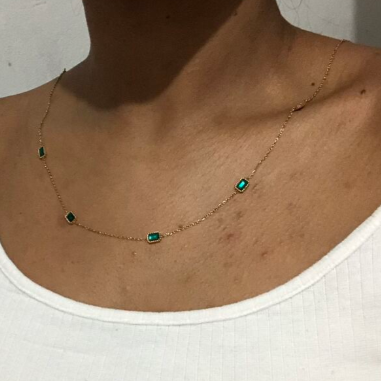 A gold chain necklace with five small rectangular green emerald-cut stones, shown worn on a person's neck above a white top.