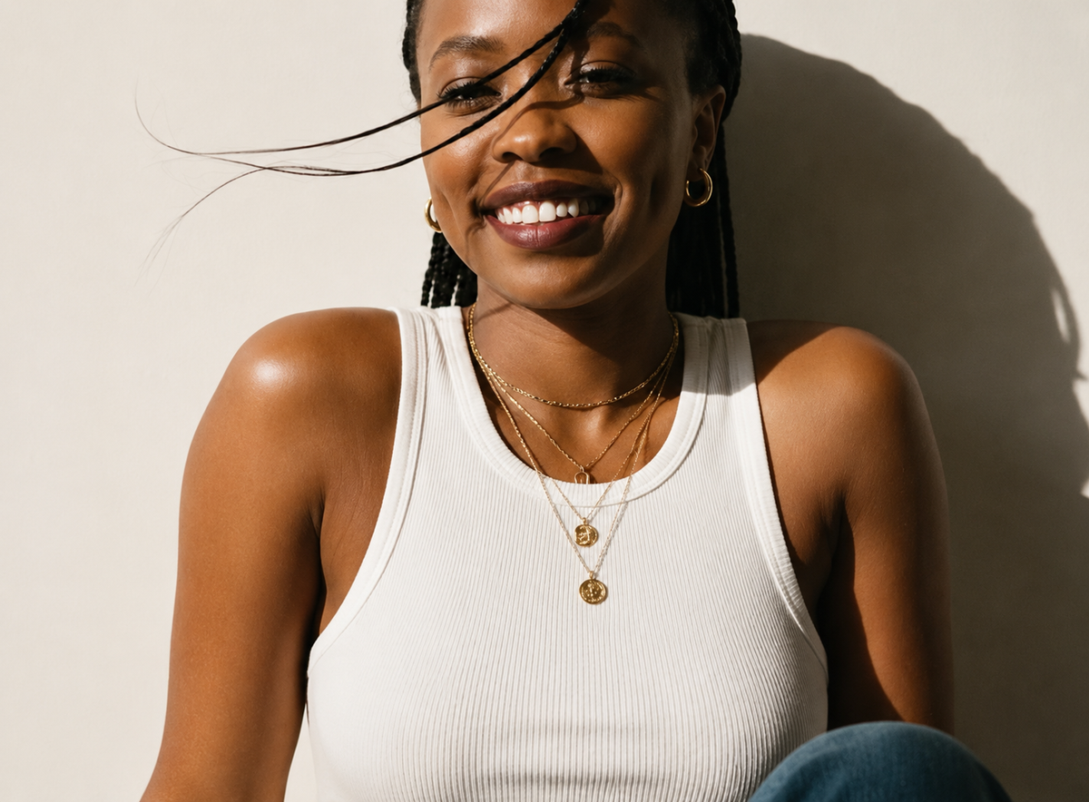 A smiling woman with braided hair wearing layered gold necklaces and hoop earrings, posing in a white ribbed tank top against a warm beige wall with a soft shadow.
