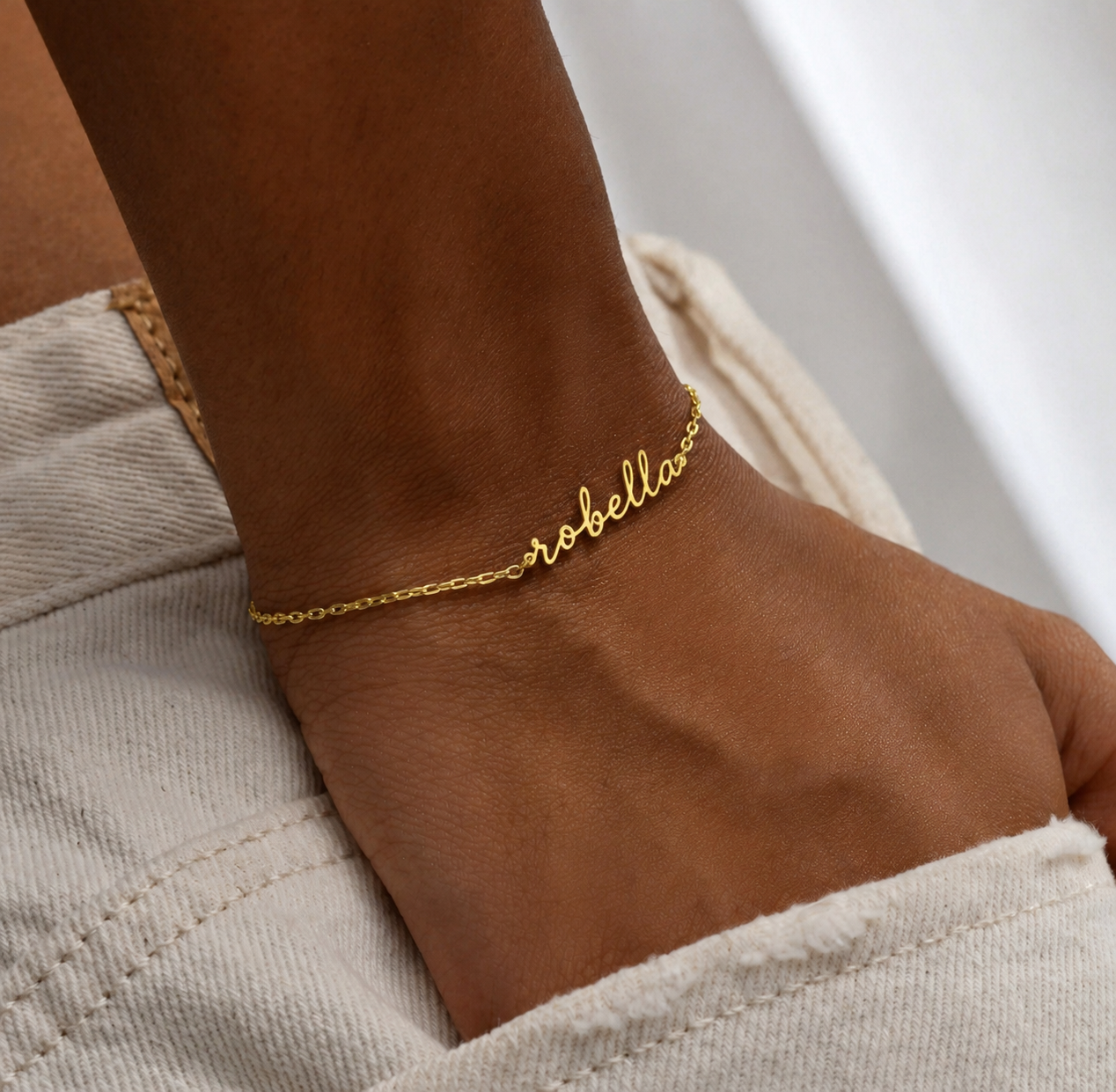 A close-up of a person's wrist wearing a delicate gold chain anklet with the name 'robella' written in cursive script, paired with light beige denim shorts.