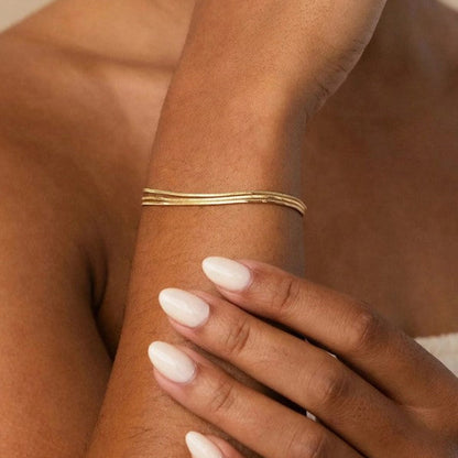 A close-up of a person's wrist wearing a thin, multi-strand gold cuff bracelet, with their hand resting on their forearm against a warm beige background.