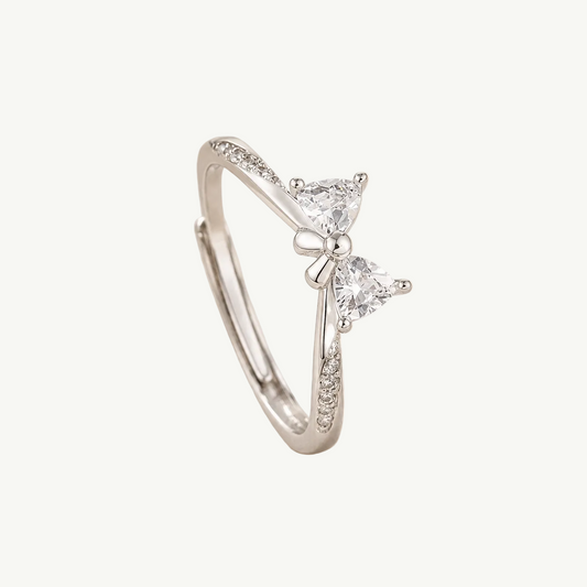 A silver-toned ring featuring two triangular clear gemstones in a bow-like setting, with small pavé accents along the band, photographed on soft cream fabric.