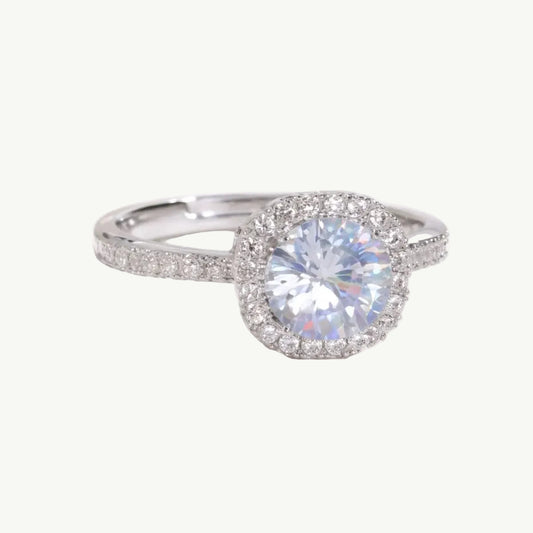 A silver halo engagement ring with a large round brilliant-cut center stone and smaller accent stones along the band, displayed on a soft white satin fabric.