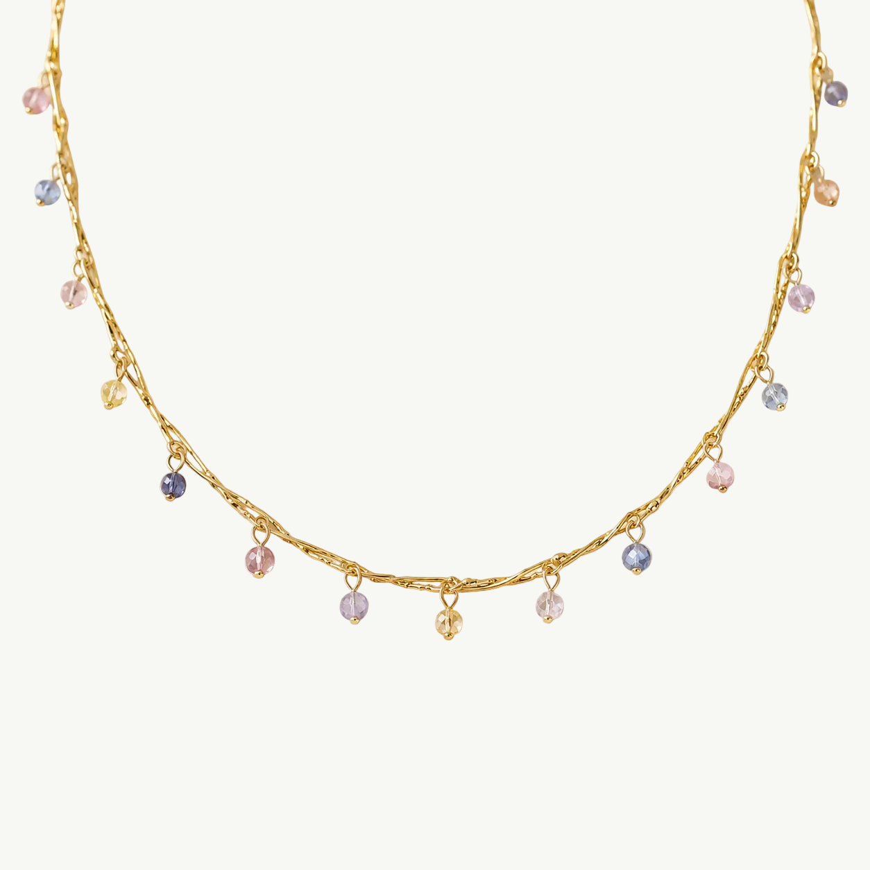 A delicate gold chain necklace with small dangling charms in pastel pink, blue, and lavender, displayed on a warm beige background.