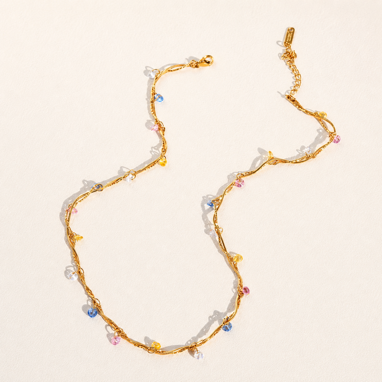 A gold chain necklace with small, colorful faceted beads in pink, blue, yellow, and white, arranged on a light beige background.