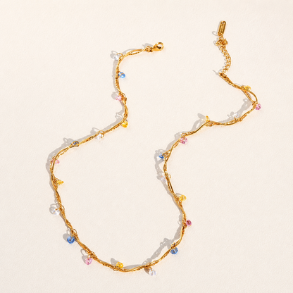A gold chain necklace with small, colorful faceted beads in pink, blue, yellow, and white, arranged on a light beige background.