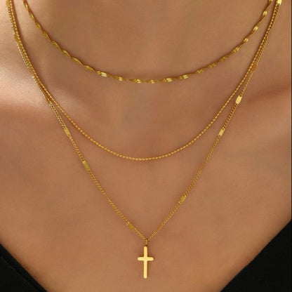 A close-up of a model's neck and chest wearing three layered gold necklaces, including a delicate chain, a textured chain, and a longer chain with a small cross pendant.