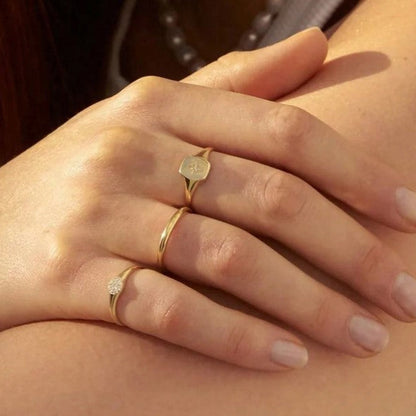 A close-up of a hand wearing three delicate gold rings, including a signet ring with a round face, a thin band, and a ring with a small clear gemstone, resting on another person's shoulder.