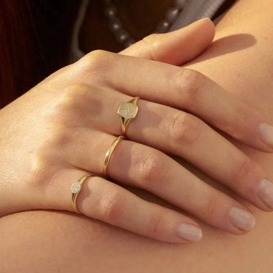 A close-up of a hand wearing three delicate gold rings, including a signet ring with a round face, a thin band, and a ring with a small clear gemstone, resting on another person's shoulder.