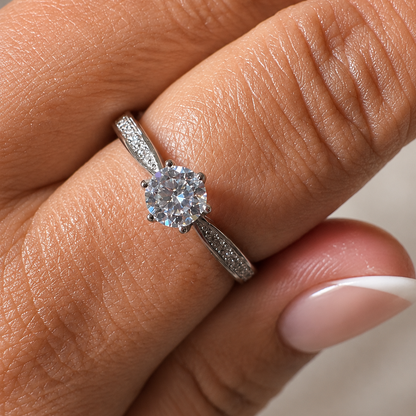 A close-up of a hand wearing a silver-toned diamond engagement ring with a round-cut center stone and pavé-set band.