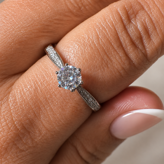 A close-up of a hand wearing a silver-toned diamond engagement ring with a round-cut center stone and pavé-set band.