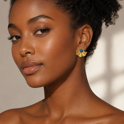 A close-up portrait of a woman wearing a gold fan-shaped earring with a blue stone accent, set against a warm beige background with soft window shadows.