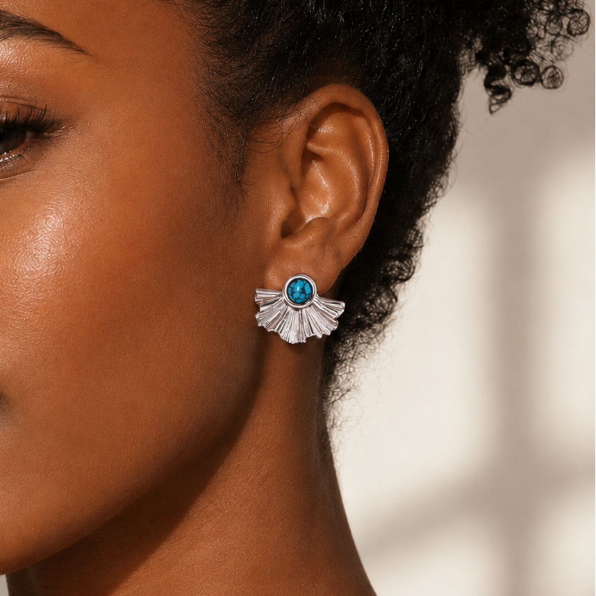 A close-up of a woman wearing a silver-toned fan-shaped stud earring with a round blue gemstone, shown against a softly lit neutral background.
