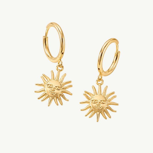 A pair of gold hoop earrings with dangling sun-face charms, displayed on a draped cream satin fabric background.