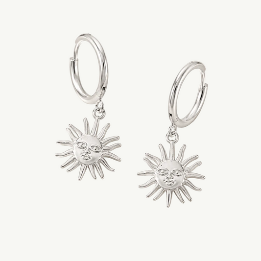 A pair of silver hoop earrings with dangling sun face charms, displayed on a soft, light beige fabric background.