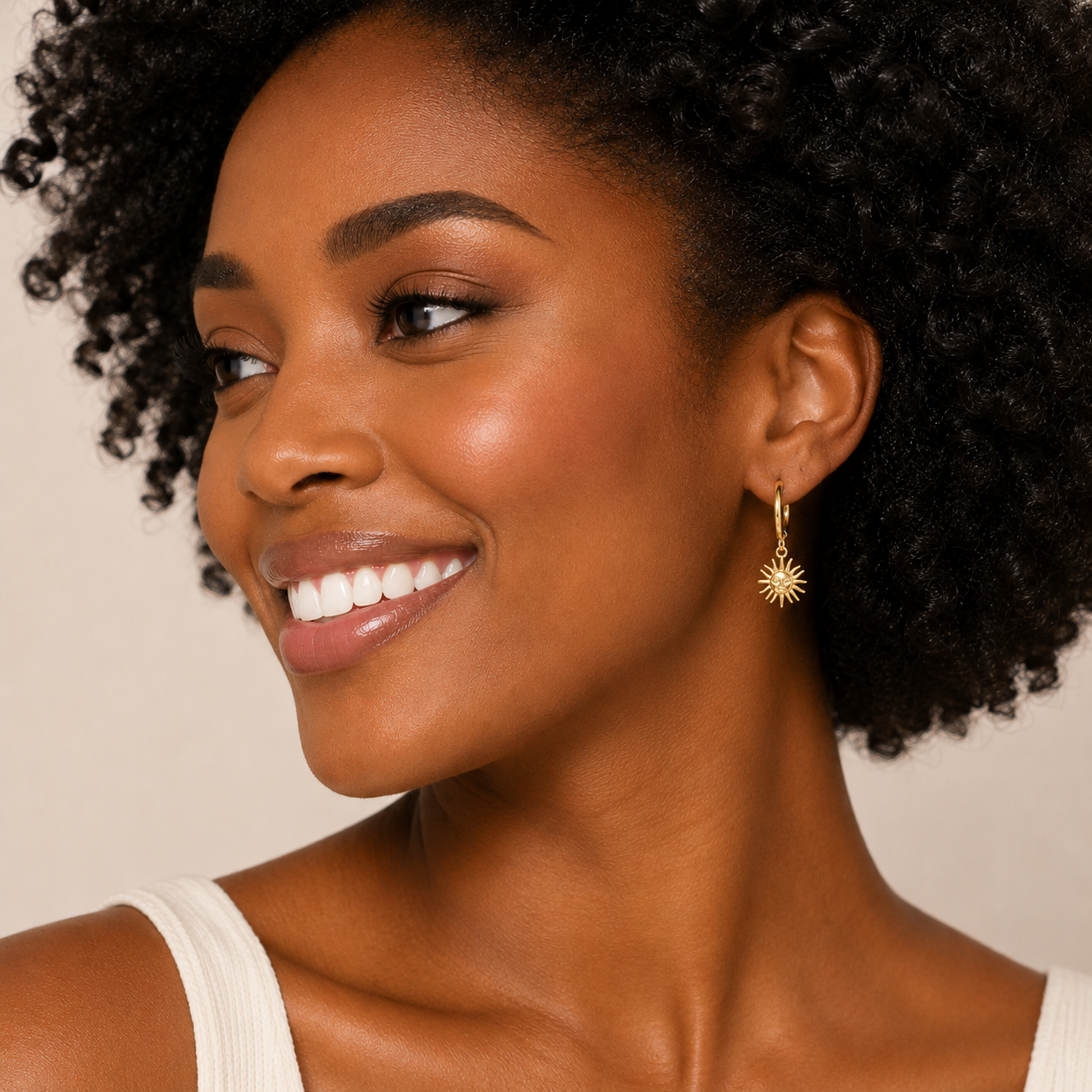 A close-up of a smiling woman with curly black hair wearing a delicate gold dangle earring with a sunburst design, set against a soft beige background.