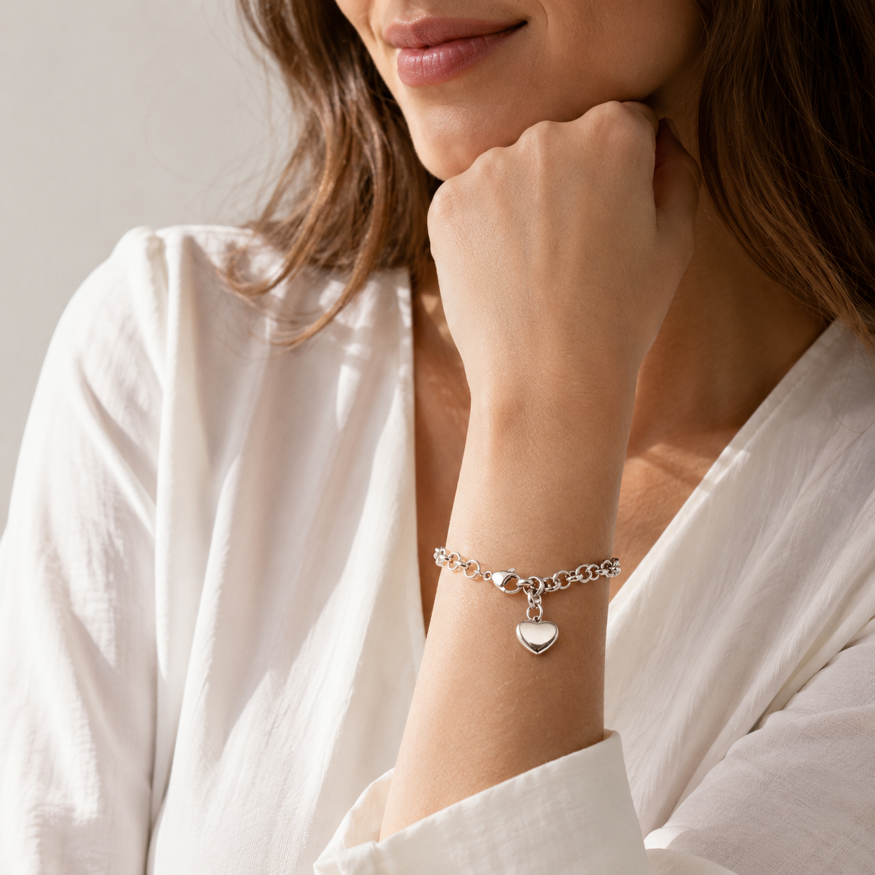 A close-up lifestyle shot of a woman wearing a delicate silver chain bracelet with a polished heart charm, styled with a white blouse.