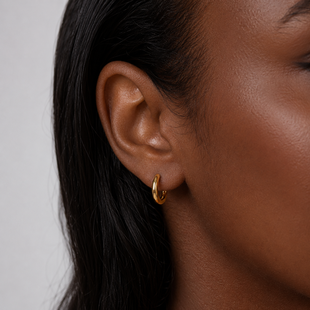 A close-up of a person's ear wearing a small gold huggie hoop earring against a light gray background.